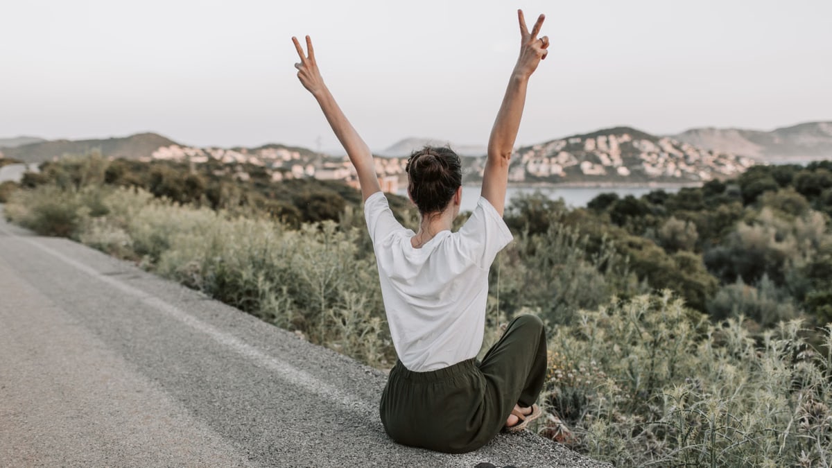 Woman on a roadside in Antalya raises peace sign, embracing summer with mountain views and tranquility.