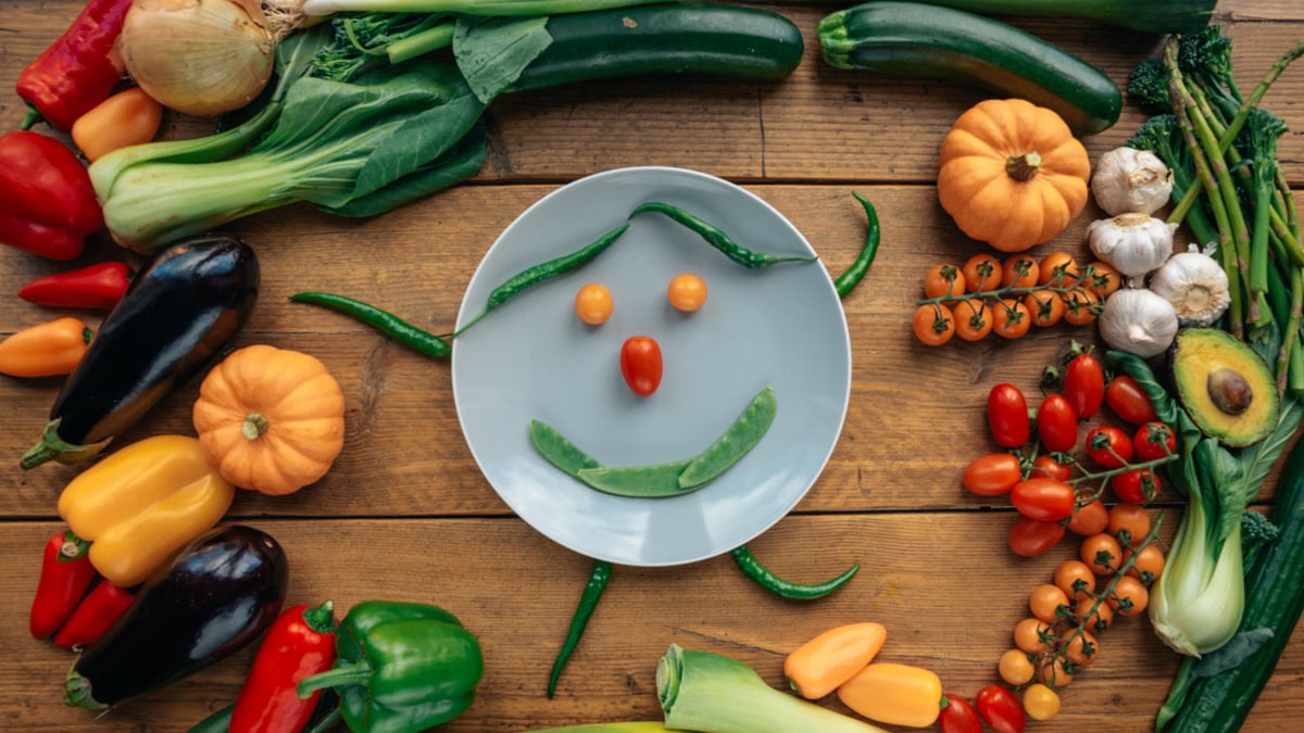 Fresh vegetables arranged creatively around a smiling plate on wooden table.